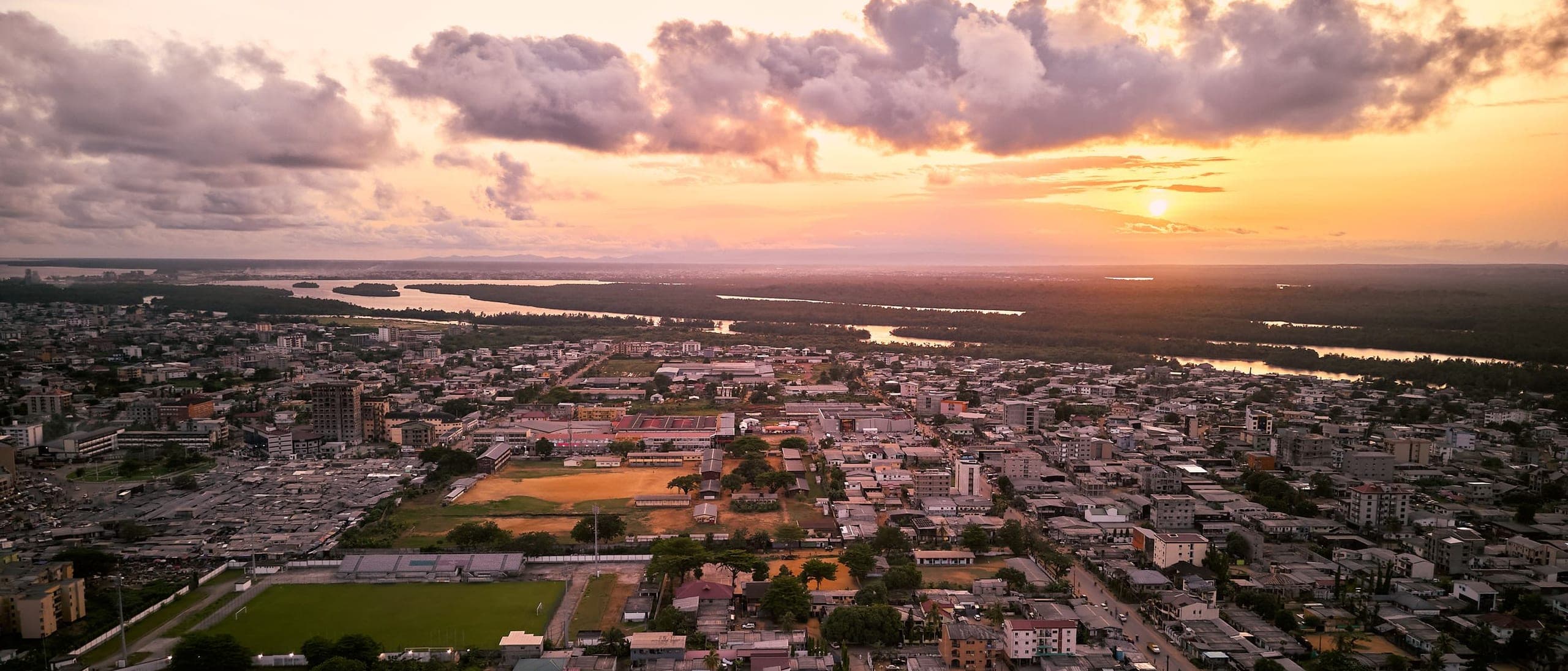 A Cameroonian city at dusk from the air — dense low-rise buildings, a river and mangrove beyond them, under a sunset sky.